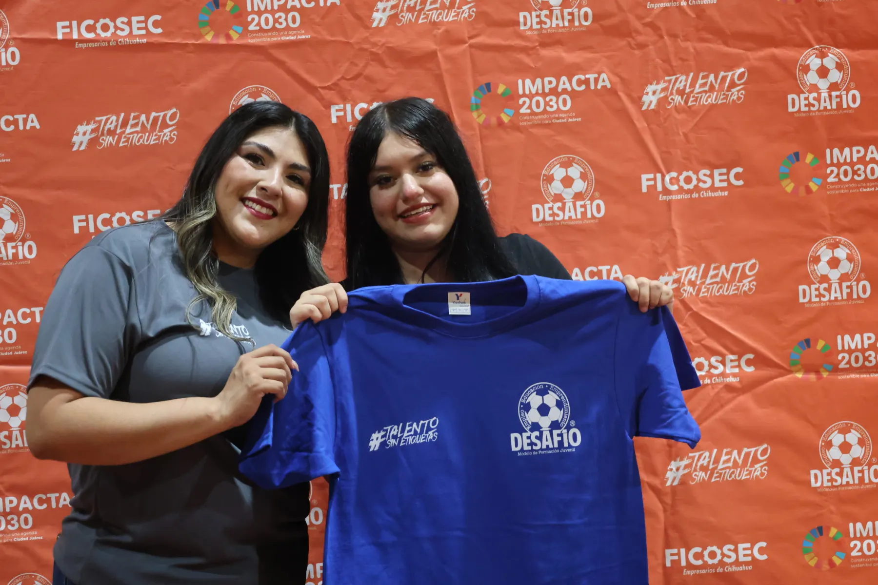 Dos jóvenes sostienen la playera de Desafío frente al fondo de FICOSEC